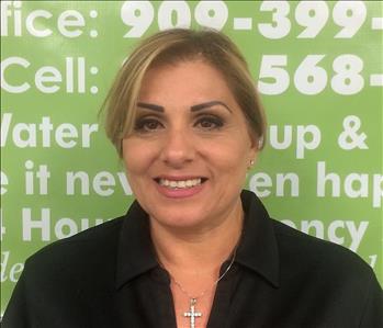 An attractive, middle-aged woman with blonde hair pulled back wearing a silver cross necklace smiling in front of a banner.