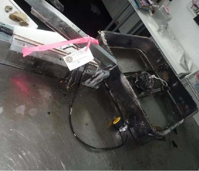 A square fan burned with the blades missing on top of a stainless steel table.