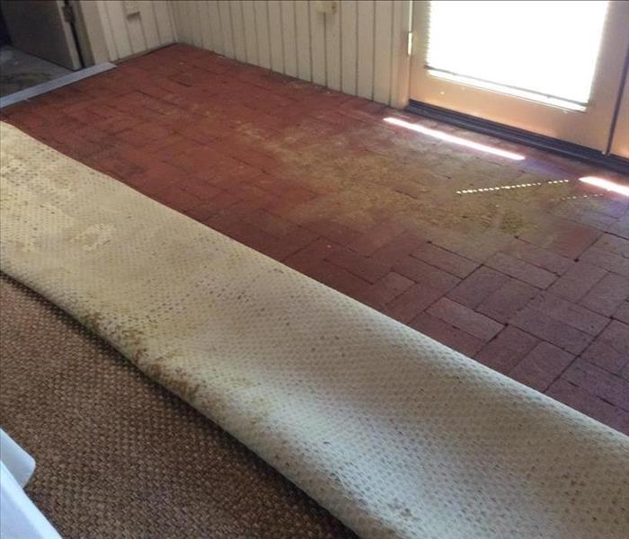 a brown carpet neatly folded over to show a red brick floor with dried mud at the doorway.